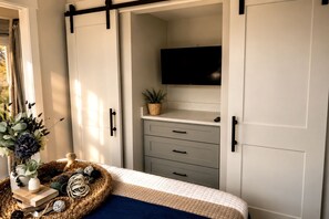 Quarto com cama, mesa de cabeceira e luminária, TV montada na parede e janela com cortinas.