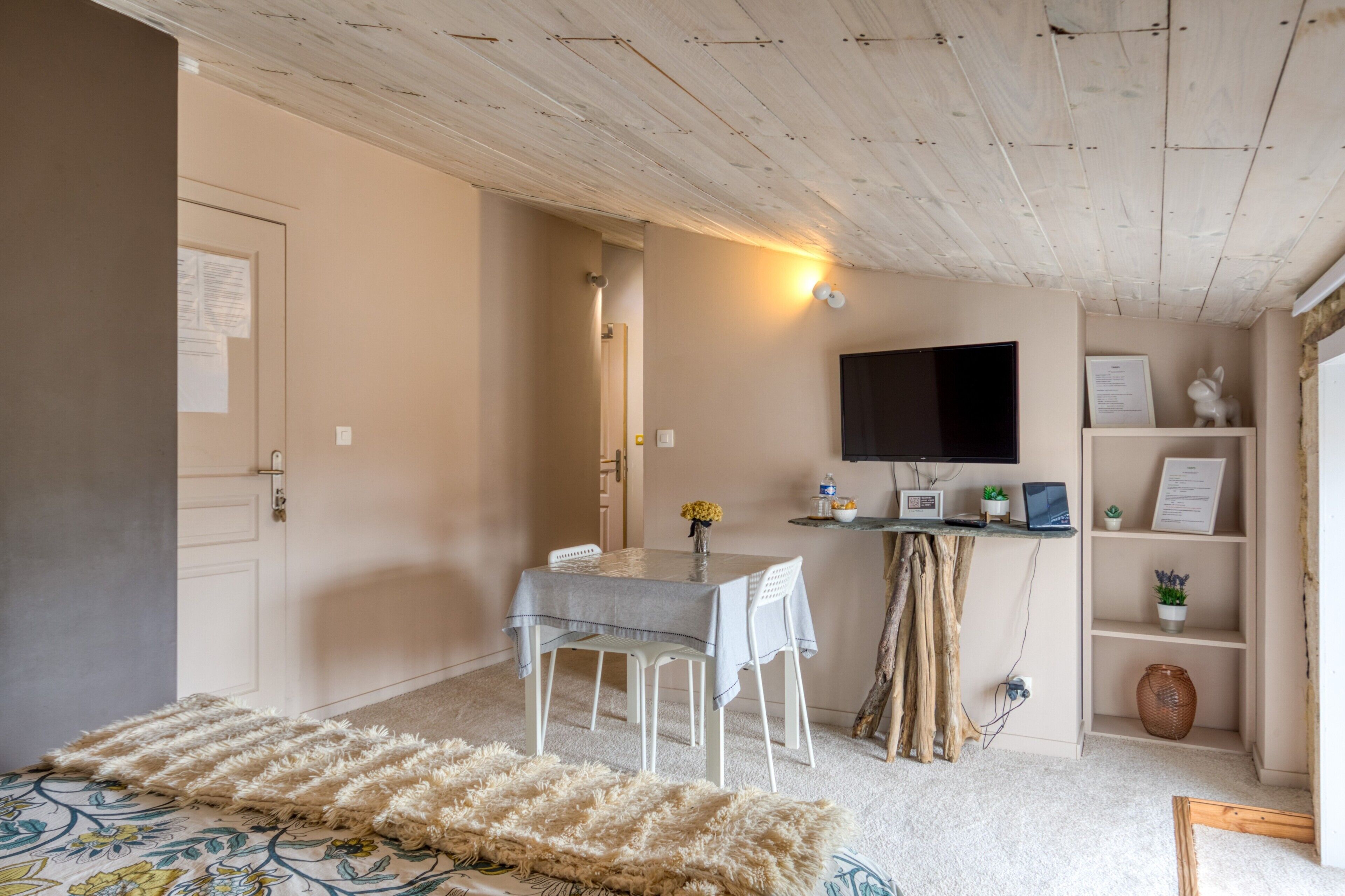 A room with a wooden ceiling, a white door, a table with chairs, a television, and a shelf with decorative items.