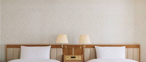 A hotel room with two beds, a wooden headboard, a nightstand with a digital clock, and lamps on the headboard.