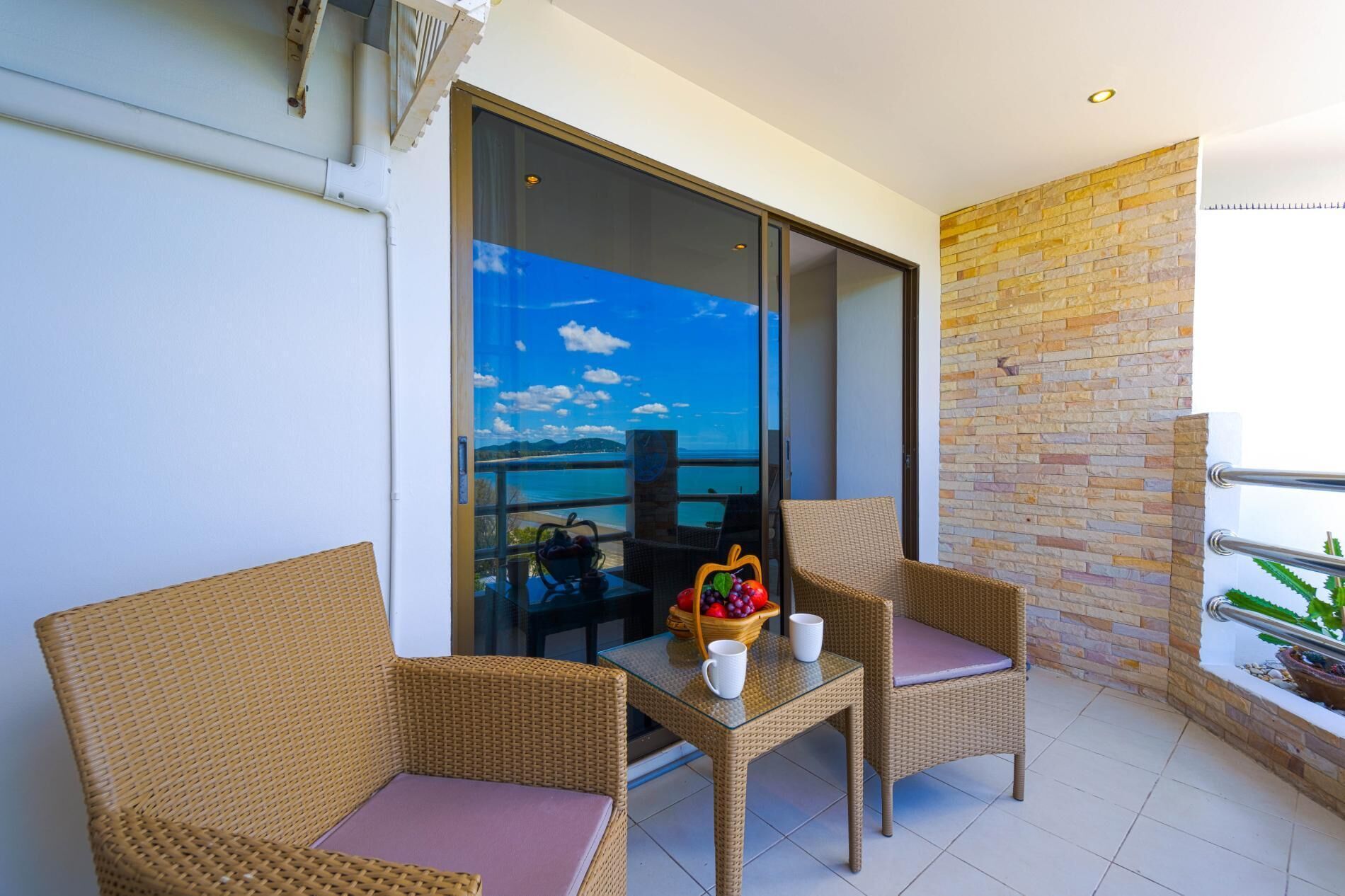 A balcony with wicker chairs, a glass table with a fruit bowl and cups, and a view of the sea.
