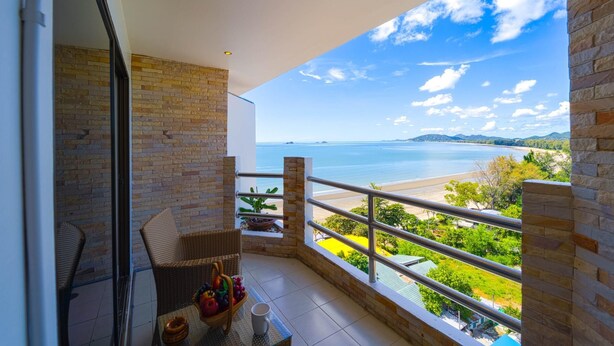 A balcony with a wicker chair, a table with fruits and a mug, and a view of the beach and ocean. - Beachfront Studio Condo-Seaview BW1 (Hua Hin)