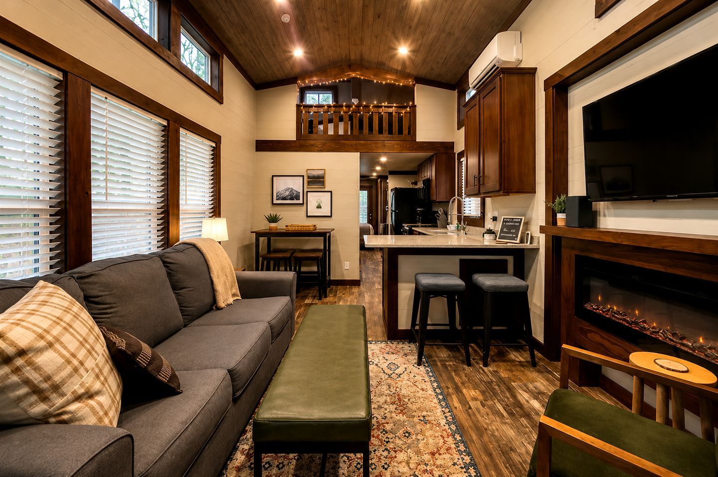 A cozy living room with a sofa, ottoman, and a dining area with a table and chairs. A kitchen area is visible with a refrigerator, stove, and a TV mounted above the counter.