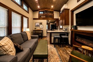 A cozy living room with a sofa, ottoman, and a dining area with a table and chairs. A kitchen area is visible with a refrigerator, stove, and a TV mounted above the counter. - Gorgeous Cabin with Hot-tub and Games in Monteagle, Tennessee (Monteagle)