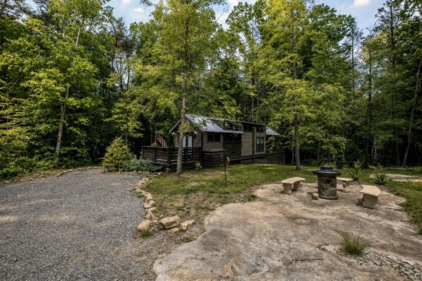 A rustic cabin surrounded by dense forest, with a gravel driveway leading to a stone patio area.