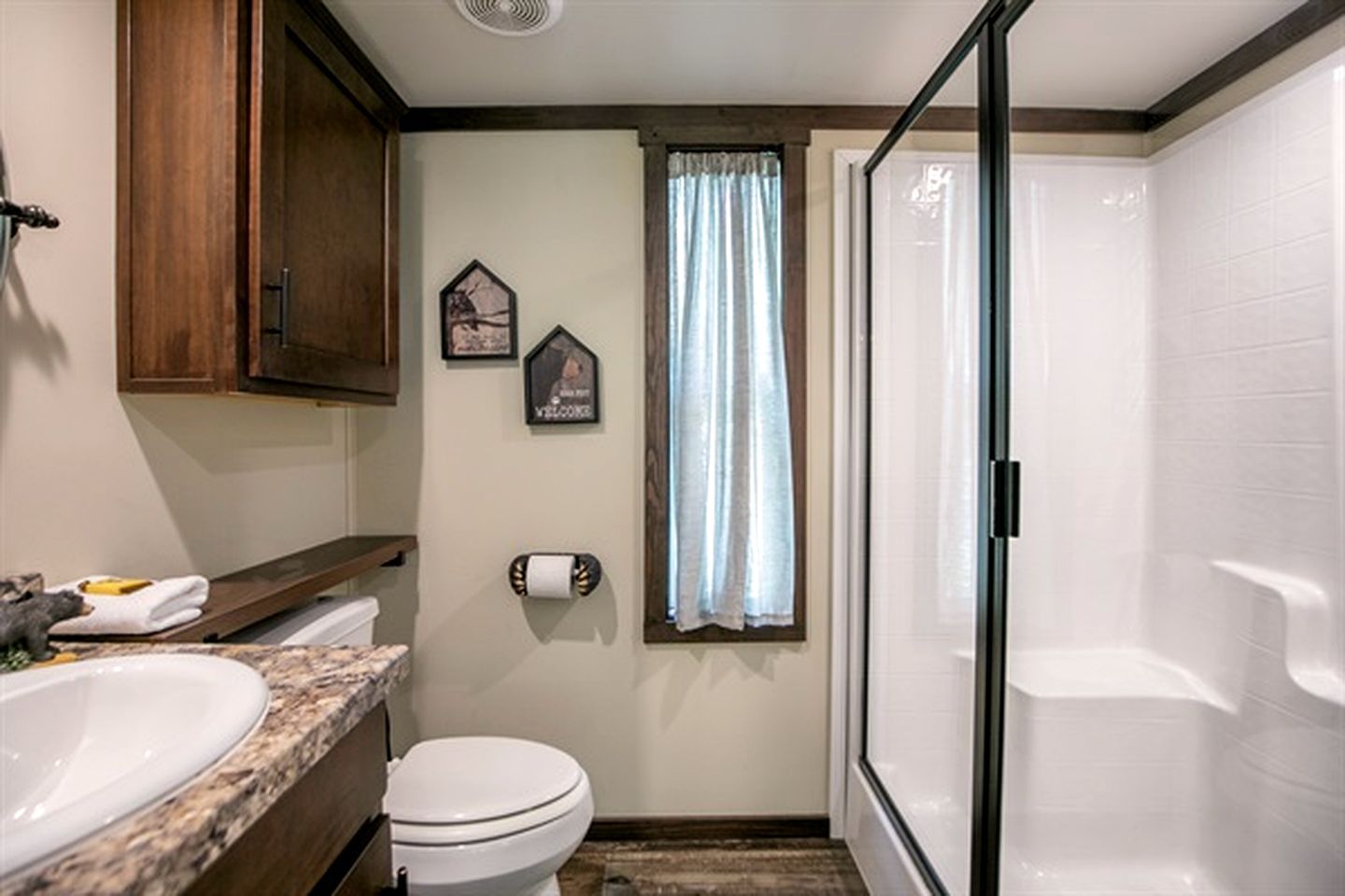 A compact bathroom with a toilet, sink, and shower. There are wooden cabinets and a window with curtains.