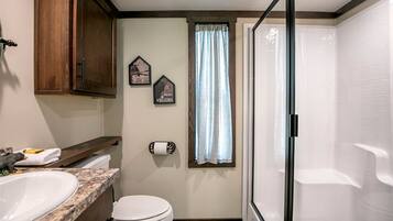 A compact bathroom with a toilet, sink, and shower. There are wooden cabinets and a window with curtains.
