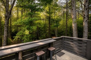 Una terraza de madera con barandilla, dos taburetes y un banco con vistas a un bosque.