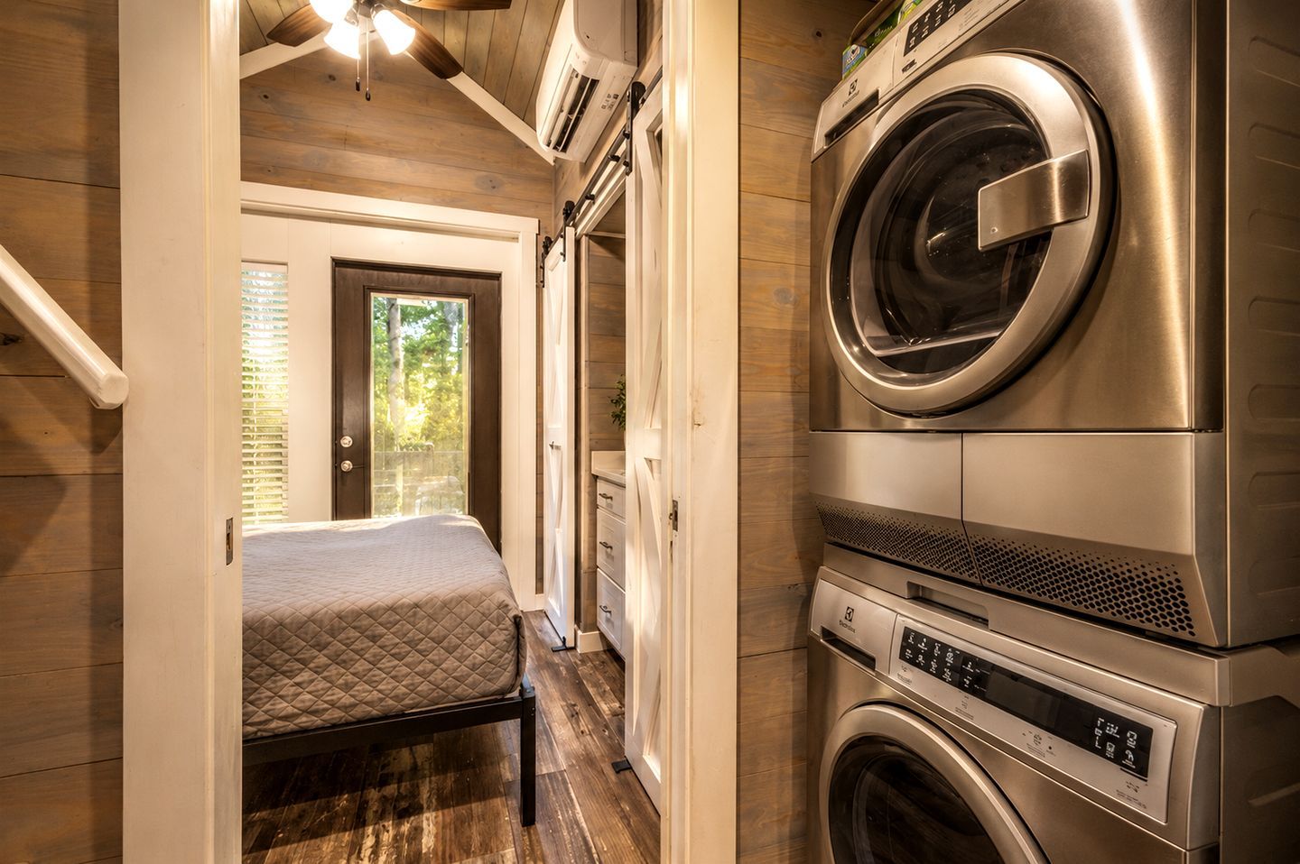 A compact laundry room with a washing machine and dryer, a bed with a quilted mattress, and a door leading to a balcony with a view of trees.