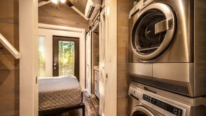 A compact laundry room with a washing machine and dryer, a bed with a quilted mattress, and a door leading to a balcony with a view of trees. - Tranquil Cabin with Fire Pit and Panoramic Lake Views in Tennessee (Tracy City)