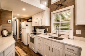 A modern kitchen with white cabinetry, a stainless steel sink, a built-in oven, and a window with blinds. - Tranquil Cabin with Fire Pit and Panoramic Lake Views in Tennessee (Tracy City)