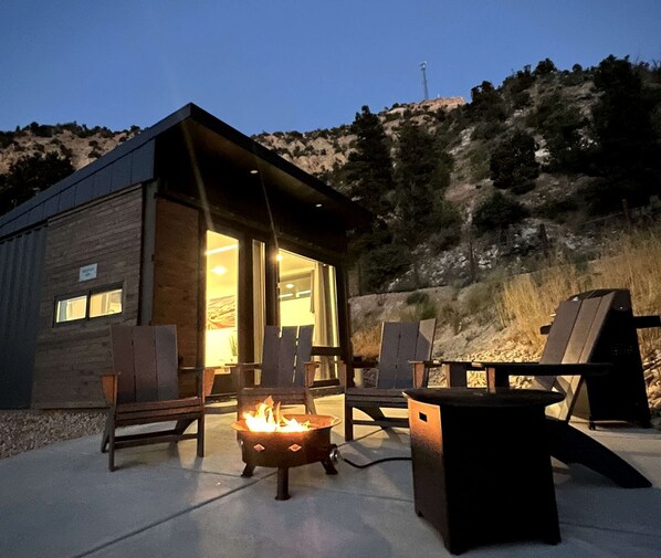 Un área de estar al aire libre acogedora con una chimenea, sillas de madera y un edificio pequeño con puerta de cristal.