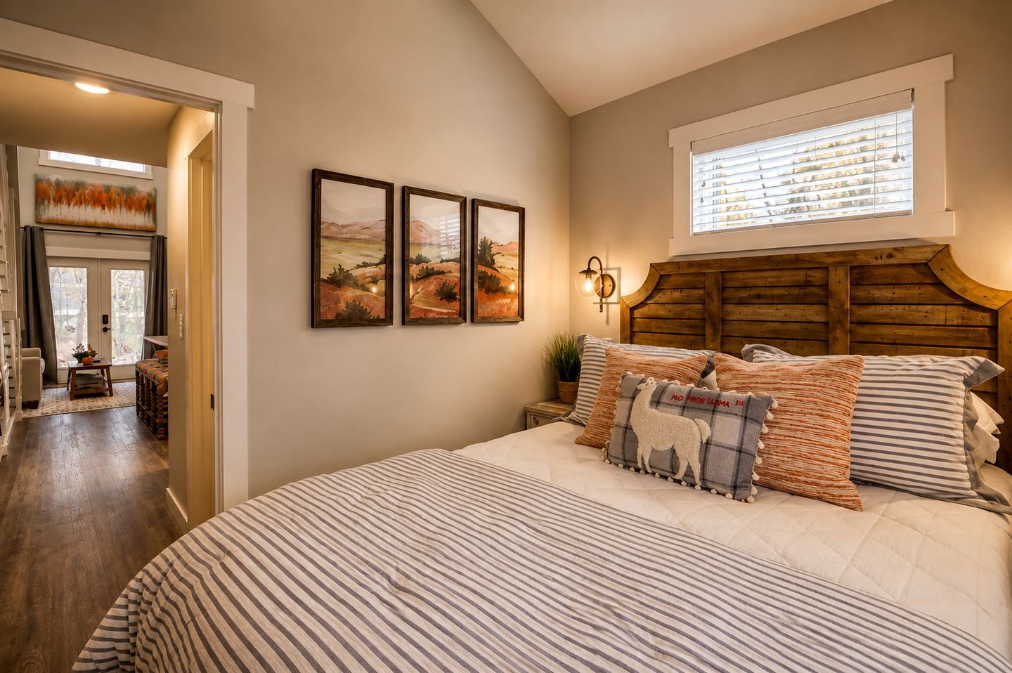 A bedroom with a large bed, a wooden headboard, and a window with blinds. There are three framed pictures on the wall.