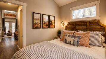 A bedroom with a large bed, a wooden headboard, and a window with blinds. There are three framed pictures on the wall.