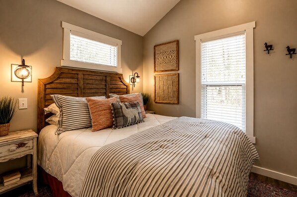 A bedroom with a wooden headboard, a large bed with striped bedding, two wall-mounted lamps, and two windows with blinds.