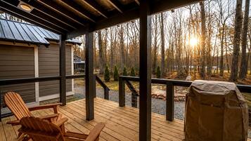 Una terraza de madera con sillas Adirondack, una parrilla y vista a un bosque.