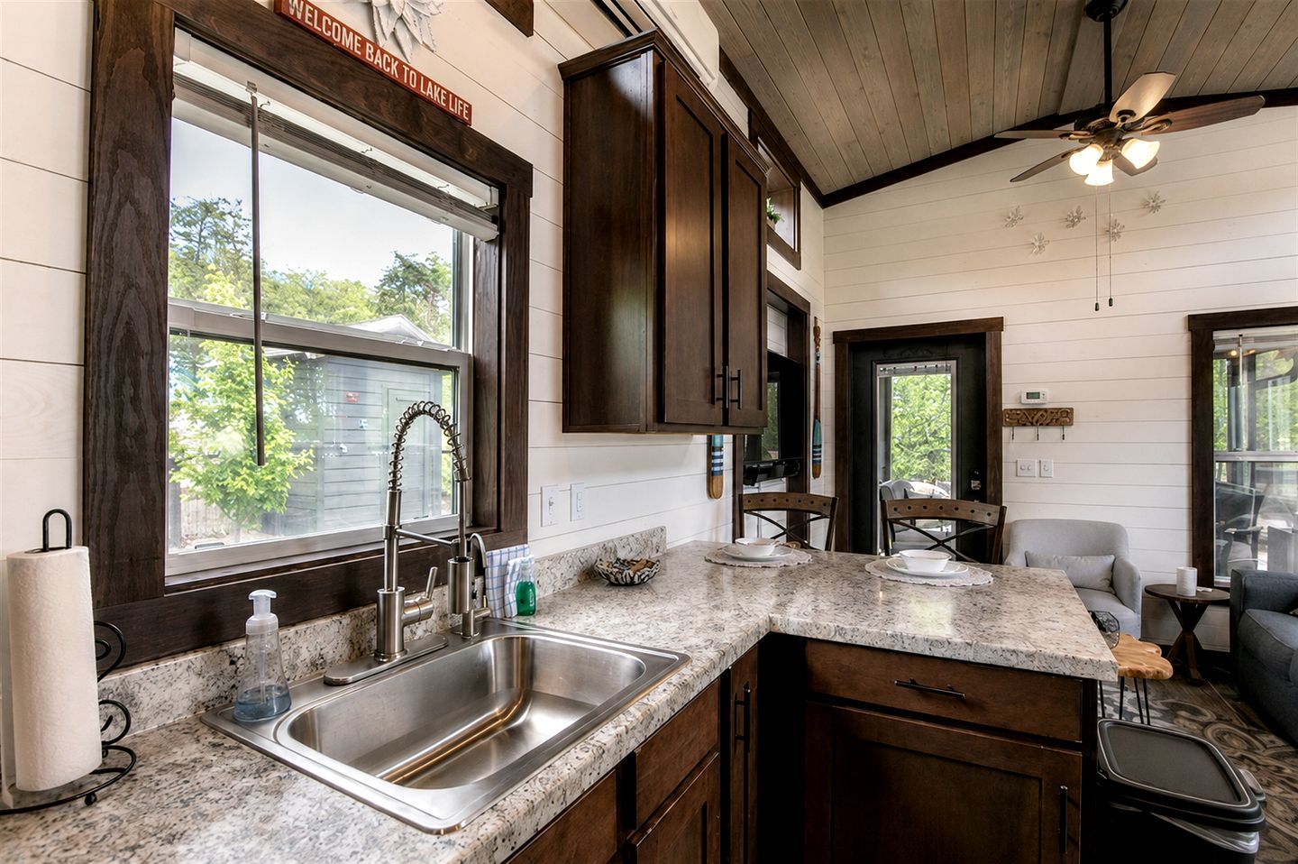 Una cocina con encimera de granito, pileta de acero inoxidable, armarios de madera y ventilador de techo.