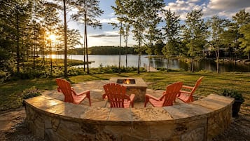 A circular stone fire pit with four chairs, surrounded by a grassy area with trees and a lake view.