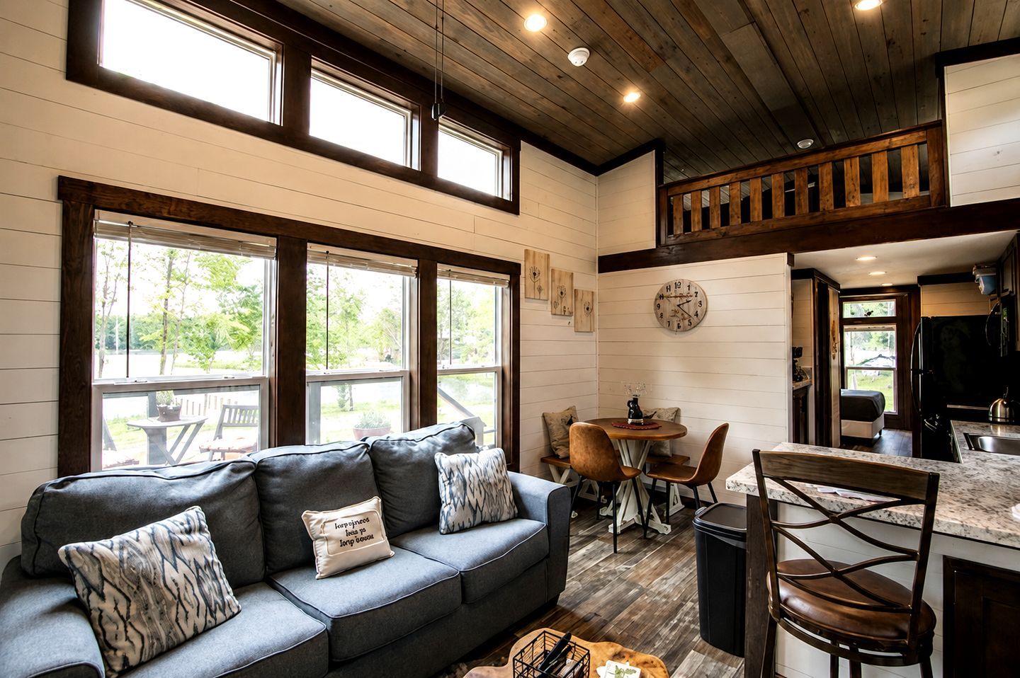 A cozy living room with a sofa, a dining table, and chairs, a kitchen area with a refrigerator, and large windows providing natural light.