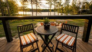 A wooden deck with a round table and two chairs, overlooking a lake with trees in the background.