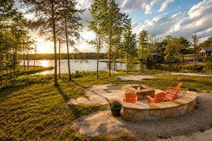 Un espace extérieur aménagé au bord du lac, avec un foyer en pierre et des chaises, entouré d’arbres et offrant une vue sur le lac.