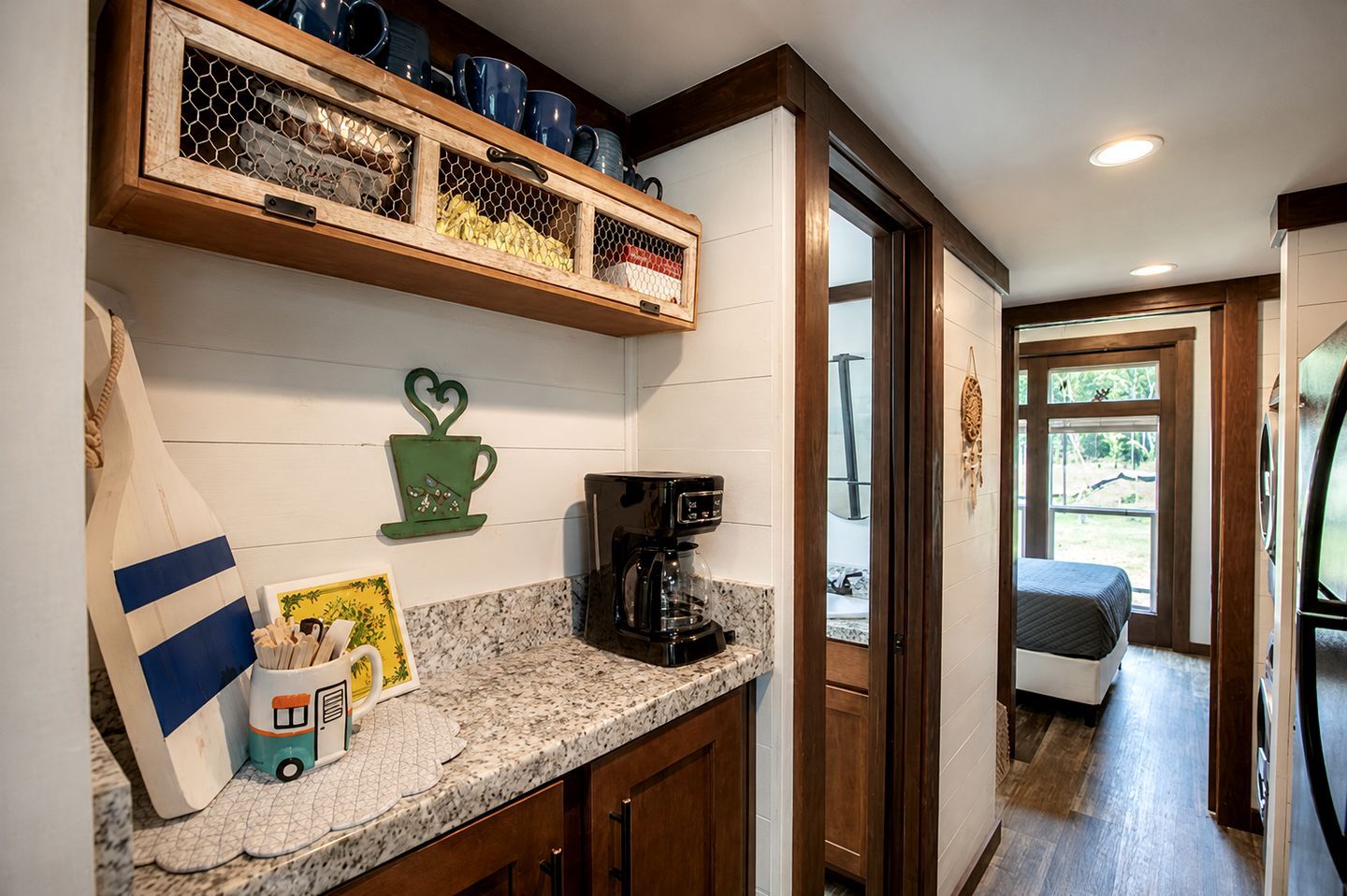 A kitchen with a coffee maker, a shelf with dishes, and a window leading to a bedroom.
