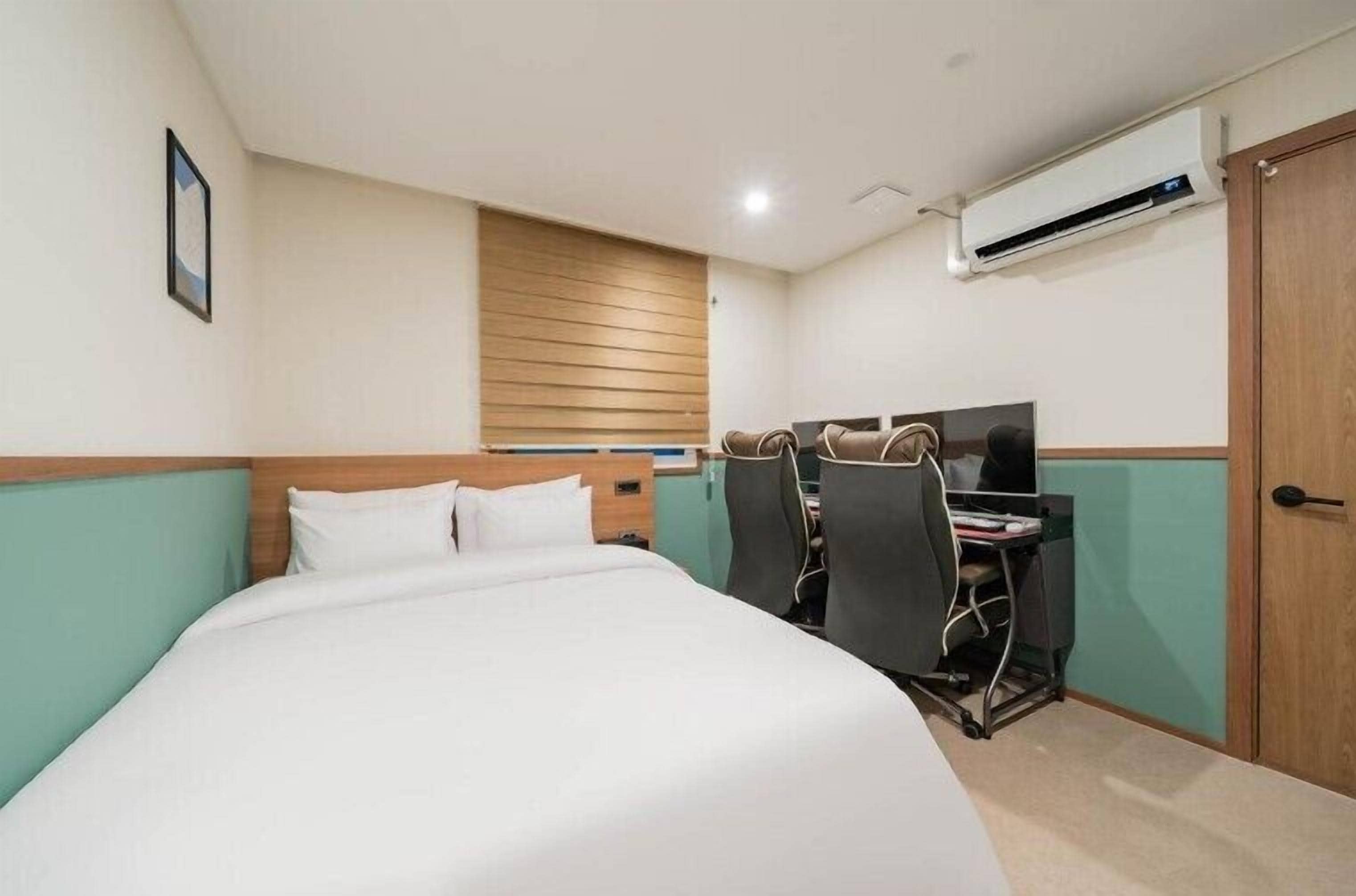 Habitación de hotel con cama, escritorio con dos sillas, equipo de aire acondicionado y ventana con listones de madera.