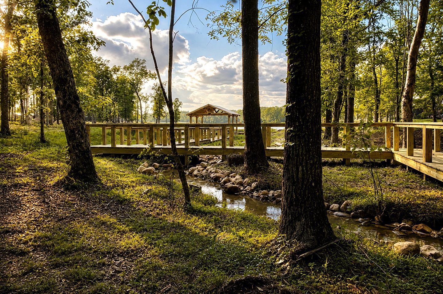 Un kiosque en bois situé dans une zone boisée, doté d’une terrasse en bois et bordé d’un petit ruisseau.