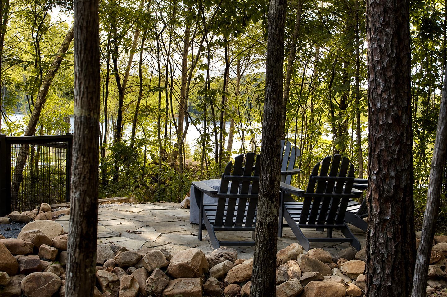 An outdoor seating area with Adirondack chairs and a stone table, surrounded by trees and a rocky landscape.