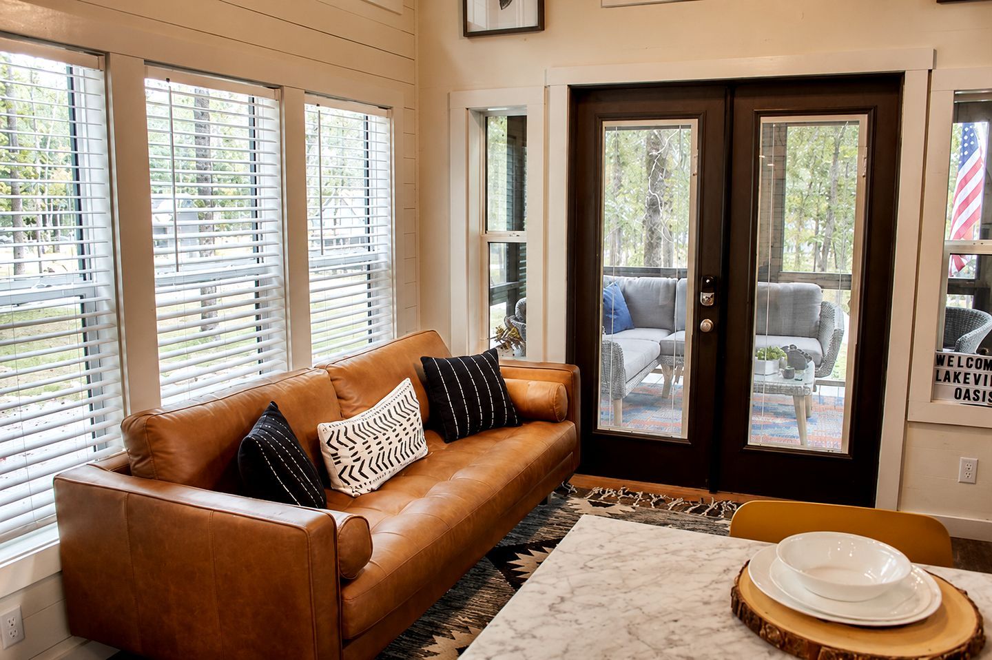 A brown leather sofa with patterned cushions, a marble table with a white plate and bowl, and a door leading to an outdoor seating area.