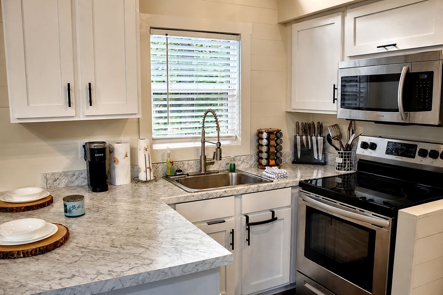 A modern kitchen with white cabinetry, a stainless steel stove, a microwave, and a window with blinds.