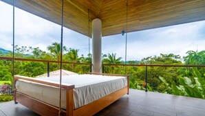 Una cama con estructura de madera y ropa de cama blanca, suspendida con cuerdas, en una habitación con vistas a exuberante vegetación y montañas.
