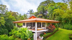 Una casa de dos pisos con un porche cubierto, rodeada de exuberante follaje y un césped cuidado.