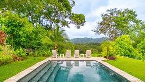 Una piscina rectangular con agua cristalina azul, rodeada de exuberante vegetación y árboles. Hay sillas de descanso blancas alineadas a lo largo del borde de la piscina.