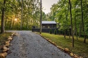 Une cabane au milieu d’une forêt, accessible par un chemin de gravier, et entourée d’arbres.