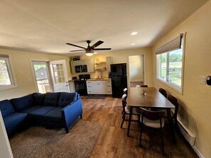 A compact living space with a kitchenette, a dining area, and a blue sectional sofa. - Beautiful Cottage Nestled Amidst Trees in Wellston, Michigan (Wellston)
