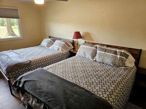 A room with two beds, a window with blinds, a lamp on a bedside table, and a view of trees outside. - Beautiful Cottage Nestled Amidst Trees in Wellston, Michigan (Wellston)
