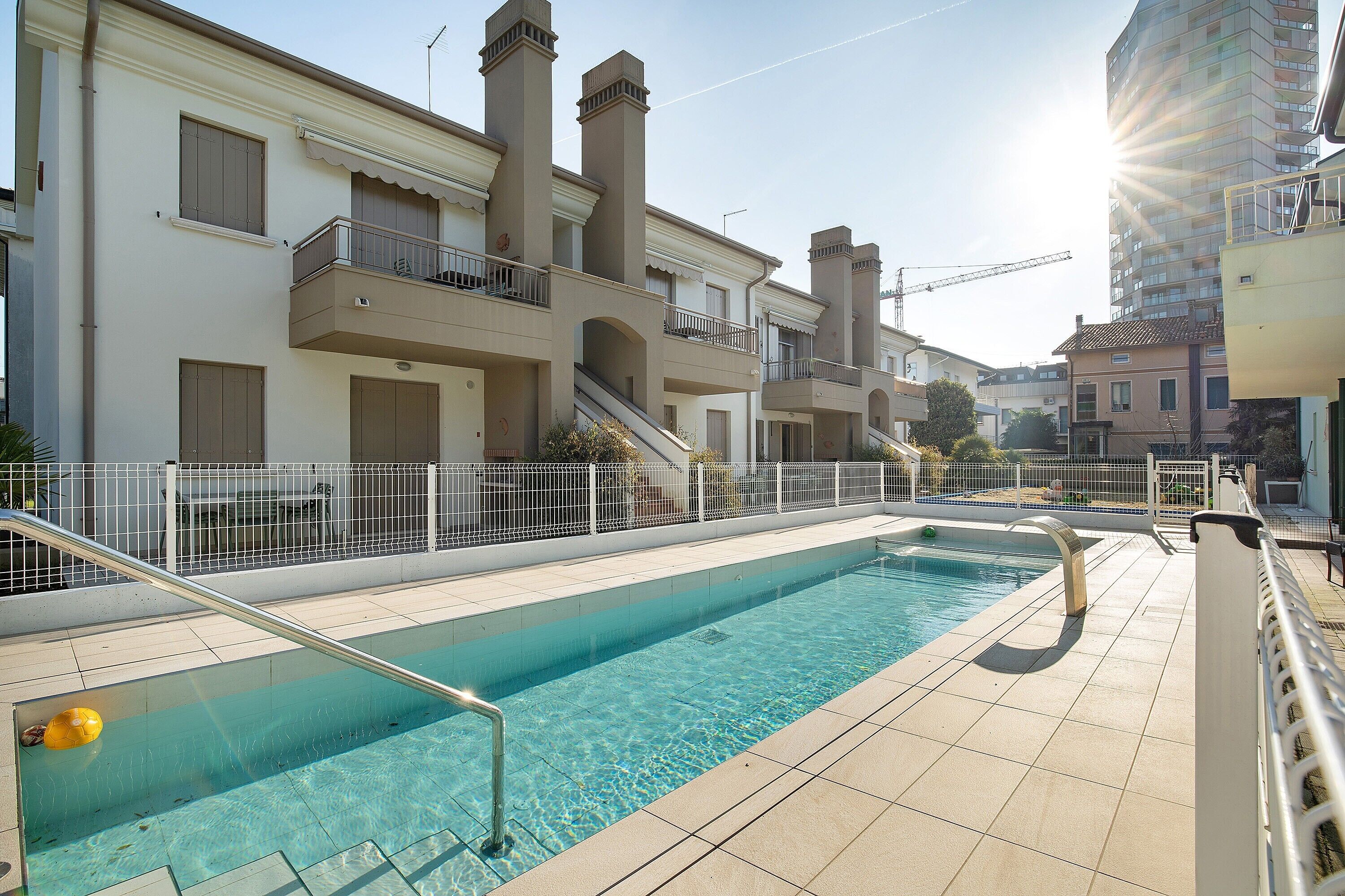A swimming pool with a metal ladder, a white fence, and a building with balconies and chimneys in the background.