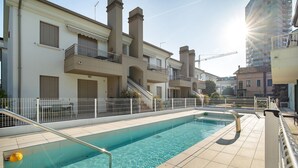 Uma piscina com uma escada de metal, um cercado branco e um edifício ao fundo, com varandas e chaminés.