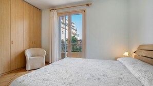 Quarto com cama, cadeira, guarda-roupa e varanda com vista para edifícios.