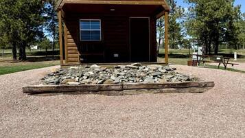 A wooden cabin with a gravel driveway and a stone fire pit in front.