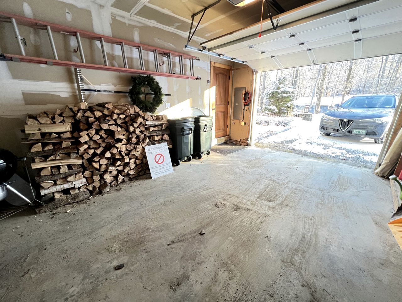 A garage with a concrete floor, stacked firewood, a sign prohibiting firewood use, a ladder, and a car parked outside.