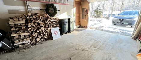 A garage with a concrete floor, stacked firewood, a sign prohibiting firewood use, a ladder, and a car parked outside.
