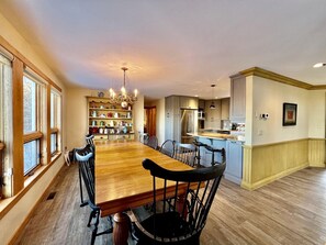 A spacious dining area with a long wooden table and chairs, a kitchen in the background, and a chandelier. - Glorious Cottage Placed in a Secluded Area in Stratton, Vermont (Stratton)