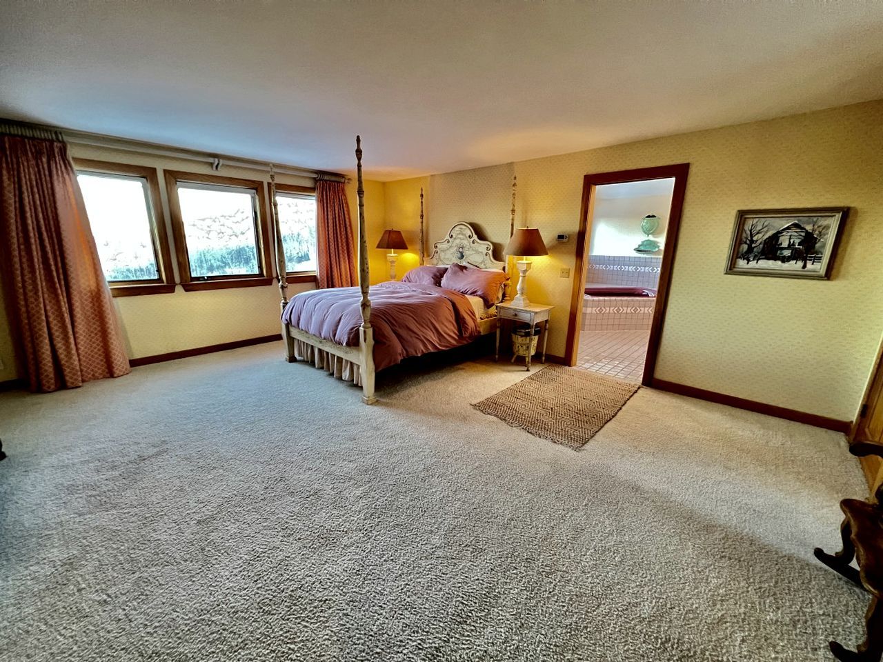 A bedroom with a four-poster bed, a small bedside table, a rug, and a door leading to another room.