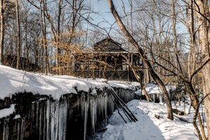 A wooden cabin with a snow-covered roof, surrounded by bare trees and icicles hanging from a stone structure. - Secluded Cabin in the Woods in Monteagle, Tennessee (Monteagle)