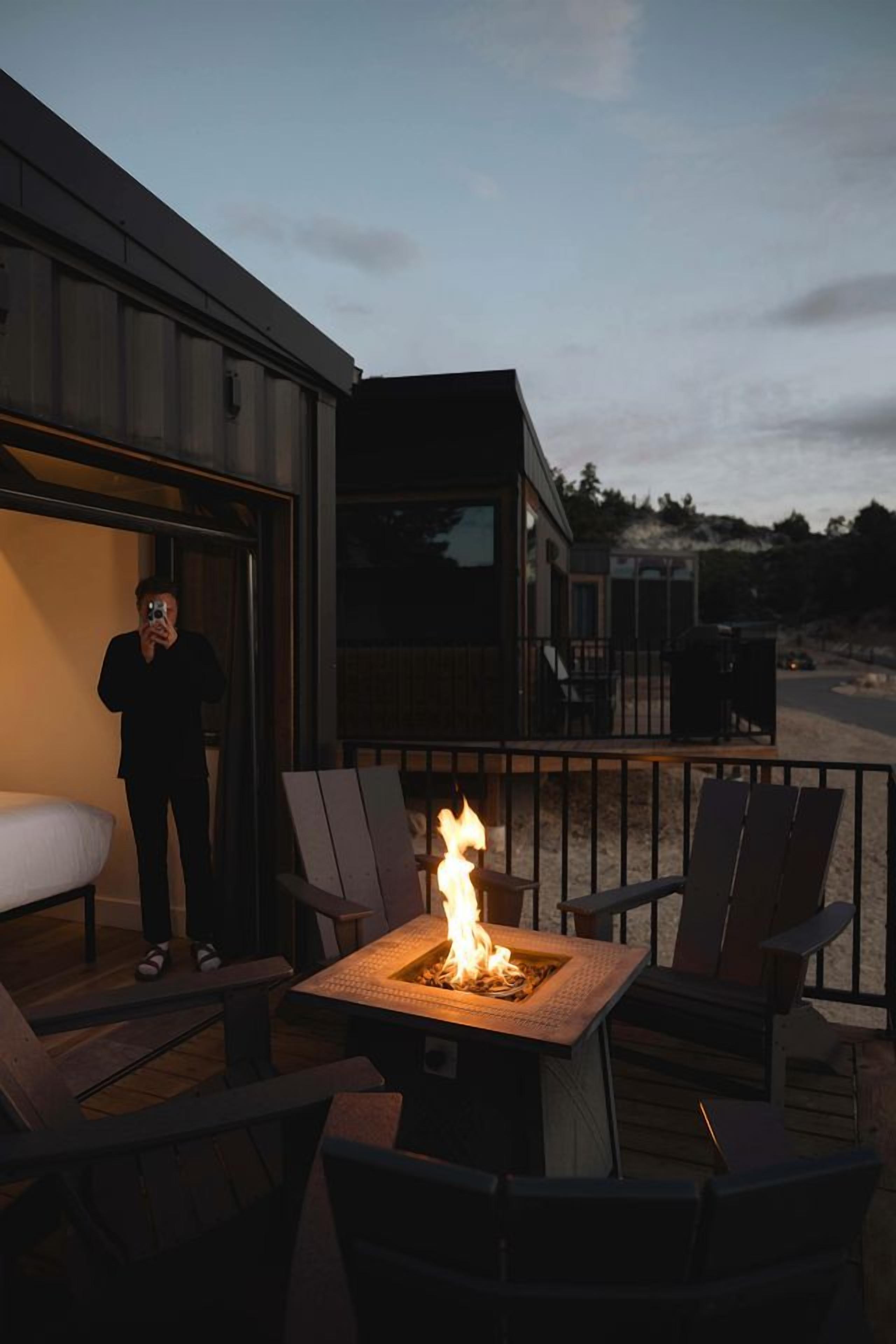 Une terrasse avec des sièges extérieurs et un foyer, une personne prenant une photo, et un bâtiment moderne en arrière-plan.