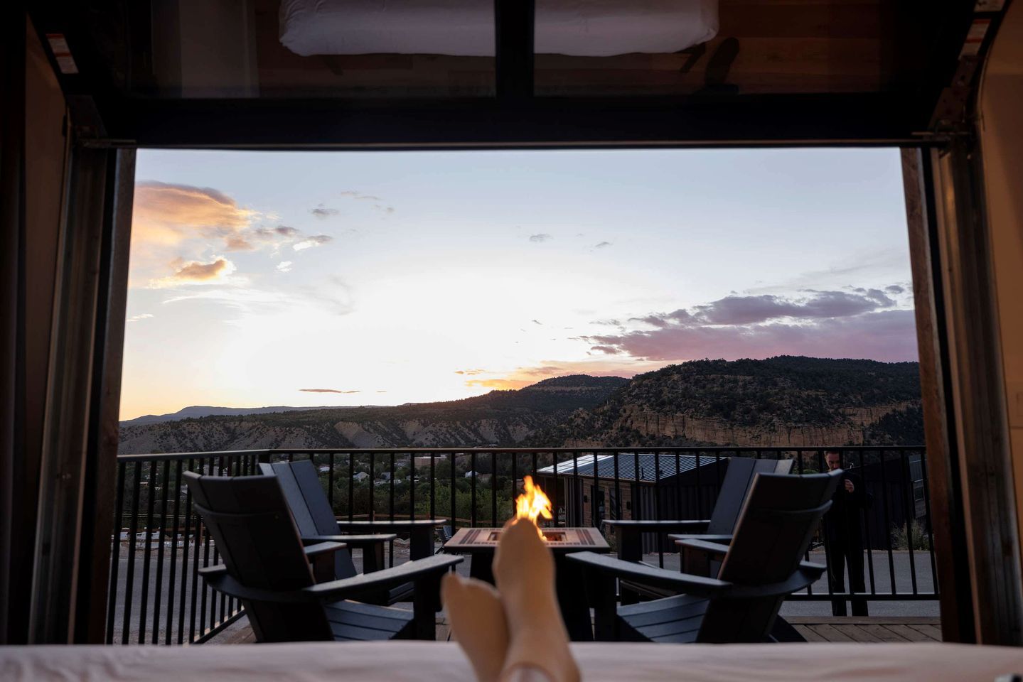 Un balcon donnant sur un paysage de montagnes au coucher du soleil, une table avec une bougie allumée et deux chaises.