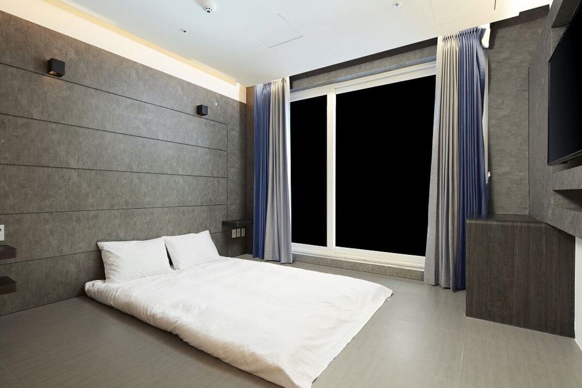 Quarto de hotel moderno com uma cama grande, televisão de tela plana e uma janela ampla com cortinas.