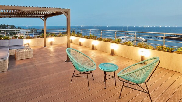 A wooden deck with wicker chairs and a table, overlooking a marina with boats and a clear view of the ocean. - Elren Hotel (Seogwipo)