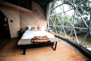 A bedroom with a bed, pillows, a blanket, a wooden bench, and a large window with a view of trees. - Quality Time in Nature: Marble Falls Luxury Glamping for Families (Marble Falls)